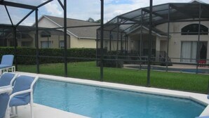 Piscina com espreguiçadeiras, cercada por um compartimento de vidro e vista para casas ao fundo.