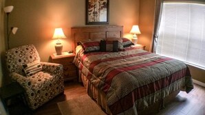A bedroom with a wooden headboard, a patterned armchair, a large bed with a quilt, two bedside lamps, and a window with blinds.