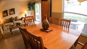 A dining area with a wooden table and chairs, a vase centerpiece, and a window with blinds. - Clubhouse 1728 Apartment 4 Bedroom (Haines City)