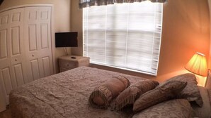 A bedroom with a large bed, a nightstand, a lamp, and a window with blinds. - Clubhouse 1728 Apartment 4 Bedroom (Haines City)