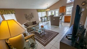 Una sala de estar con sofá, mesa de centro y lámpara. Se observa una cocina con armarios y una mesa de comedor.