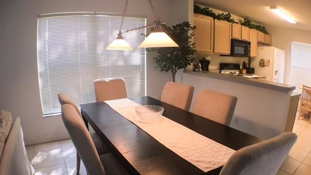 A dining area with a dark wooden table, beige chairs, and a white table runner. There is a window with blinds, a plant, and a kitchen area in the background.