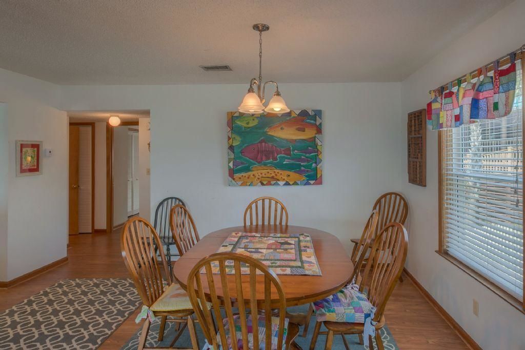 Un coin repas avec une table ronde, six chaises en bois, un tableau coloré représentant des poissons accroché au mur et une fenêtre avec des stores.