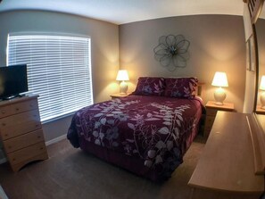 A bedroom with a bed, two bedside lamps, a dresser, a TV, and a large window with blinds. - Kokomo 2892 Apartment 3 Bedroom (Haines City)
