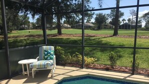 A pool area with a chair and table, surrounded by a glass enclosure with a view of a grassy yard and trees. - Mariner 1146 Apartment 4 Bedroom (Haines City)