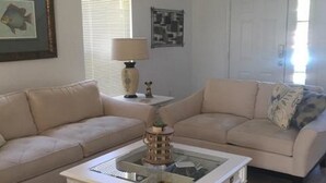 A living room with two beige sofas, a white coffee table with a decorative item, a lamp, and framed pictures on the walls. - Mariner 1146 Apartment 4 Bedroom (Haines City)