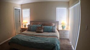 A bedroom with a large bed, two bedside tables, and two windows with blinds.
