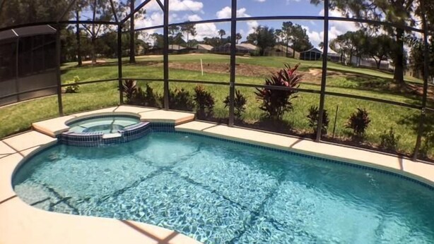 A swimming pool with a hot tub, surrounded by a fence and a view of a grassy area with trees. - Mariner Pad Apartment 4 Bedroom (Haines City)