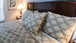 A neatly made bed with a patterned bedspread and pillows, a wooden headboard, a bedside lamp, and a framed picture on the wall. - Mariner Pad Apartment 4 Bedroom (Haines City)