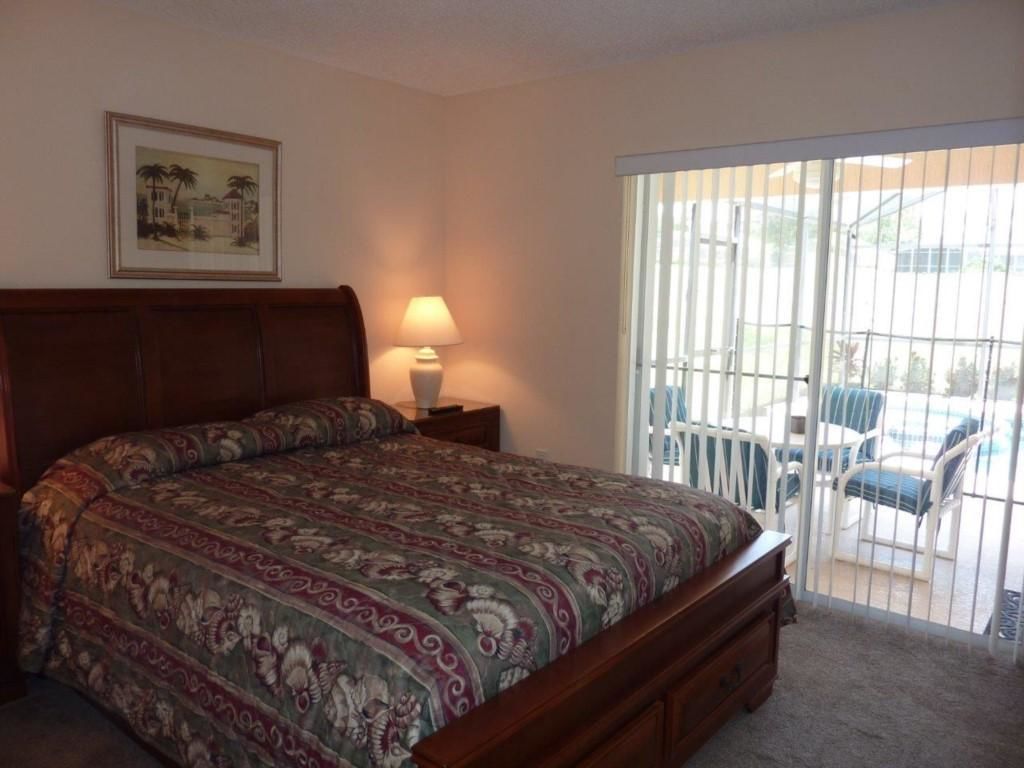 Quarto com cama, mesa de cabeceira, luminária, quadro pendurado na parede e porta de vidro corrediça que dá para área externa com cadeiras.