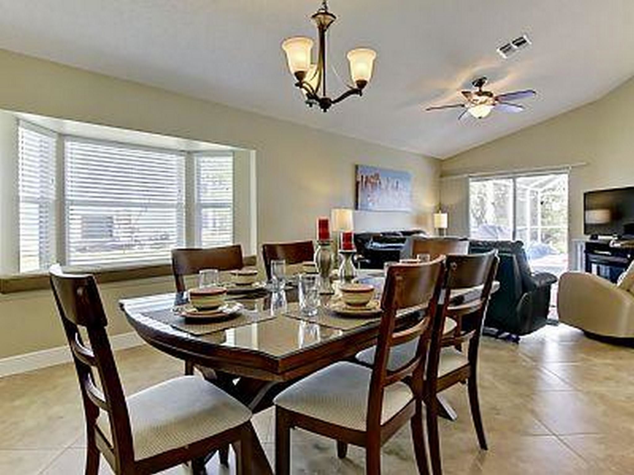 A dining area with a round table, chairs, and a chandelier. There is a television and a window with blinds in the background.
