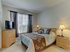 Un dormitorio con cama, dos mesitas de noche, un televisor sobre una cómoda y una ventana con cortinas.
