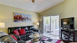 A living room with a black leather sofa, a white armchair, a glass coffee table, a flat-screen TV, and a painting on the wall.