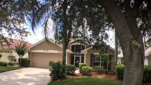 A residential house with a garage, a well-maintained lawn, and a large tree in the foreground. - Mallory Villa Villa 3 Bedroom (Haines City)
