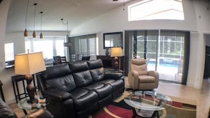 A modern living room with a black leather sofa, a beige armchair, and a glass coffee table. There are pendant lights, a lamp, and a large window with a view of the outdoors. - Mallory Villa Villa 3 Bedroom (Haines City)