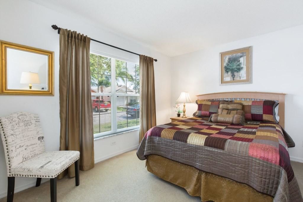 A bedroom with a large bed, a chair, a window with curtains, a bedside table with a lamp, and a framed picture on the wall.