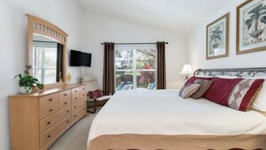 Une chambre avec un grand lit, une commode en bois, un téléviseur fixé au mur et une fenêtre avec des rideaux.