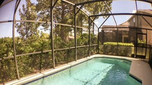 A swimming pool with a glass enclosure, surrounded by trees and a house in the background. - Hemingway Golf View Apartment 4 Bedroom (Haines City)