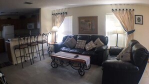 A living room with a black leather sofa, a coffee table, and a dining area with a bar stool. - Hemingway Golf View Apartment 4 Bedroom (Haines City)