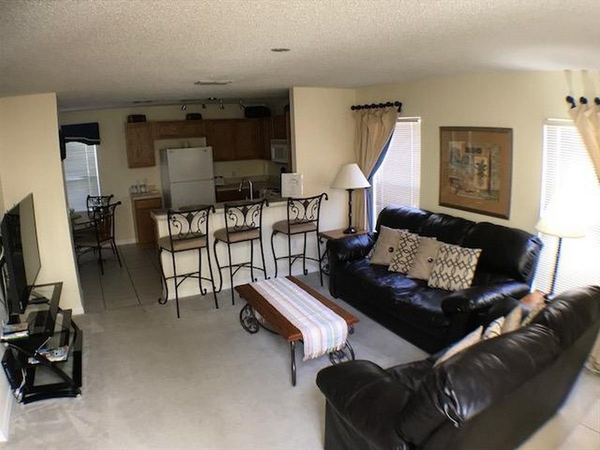 A living room with a black leather sofa, a small table, and a TV. There is a kitchen area with cabinets and a refrigerator.