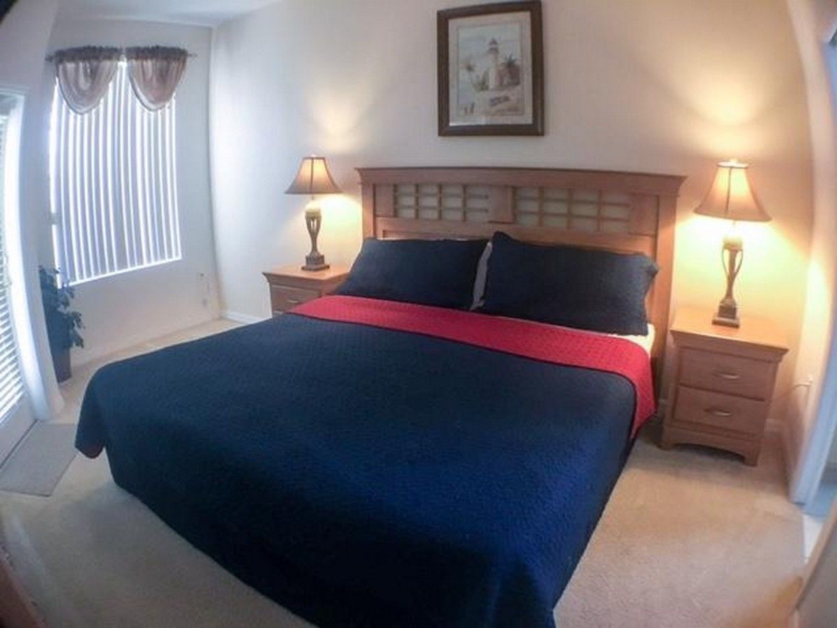 A bedroom with a large bed, two bedside tables with lamps, a window with blinds, and a framed picture on the wall.