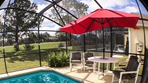A swimming pool with a red umbrella, surrounded by white chairs and a table, with a view of a grassy area and trees. - Kokomo 2749 Apartment 6 Bedroom (Haines City)