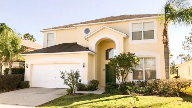 A single-story house with a garage, a small front yard with a lawn and a palm tree, and a two-car garage. - Kokomo 2749 Apartment 6 Bedroom (Haines City)