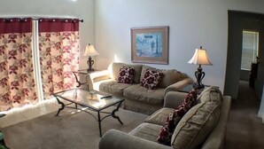 A living room with two sofas, a coffee table, and two lamps. There is a window with curtains and a framed picture on the wall. - Kokomo Golf View Apartment 4 Bedroom (Haines City)