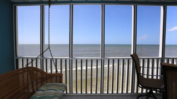 A room with a large window overlooking the beach, a wicker sofa, and a chair.