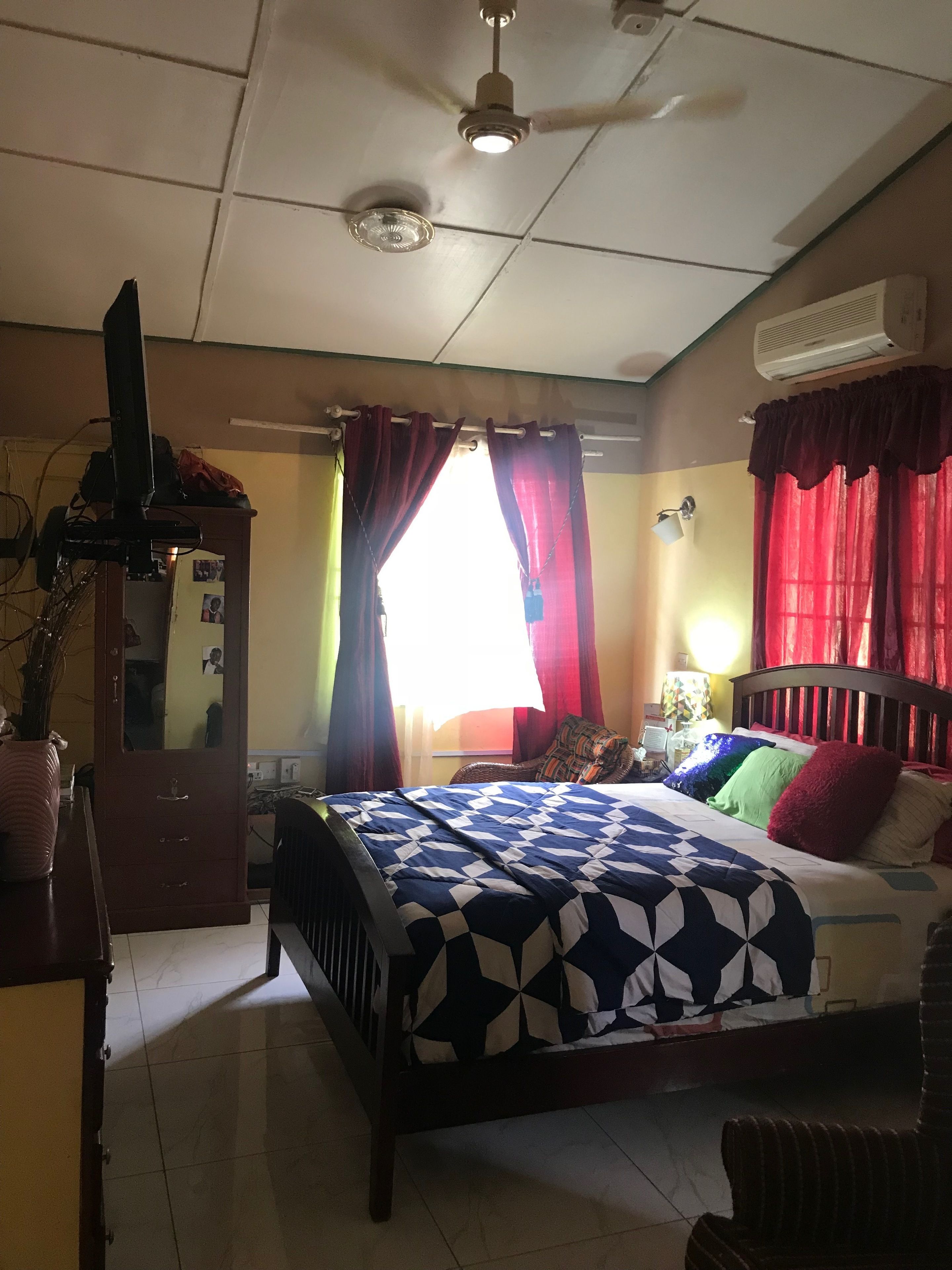 Un dormitorio con una cama, ventilador de techo, aire acondicionado, televisor y una ventana con cortinas.