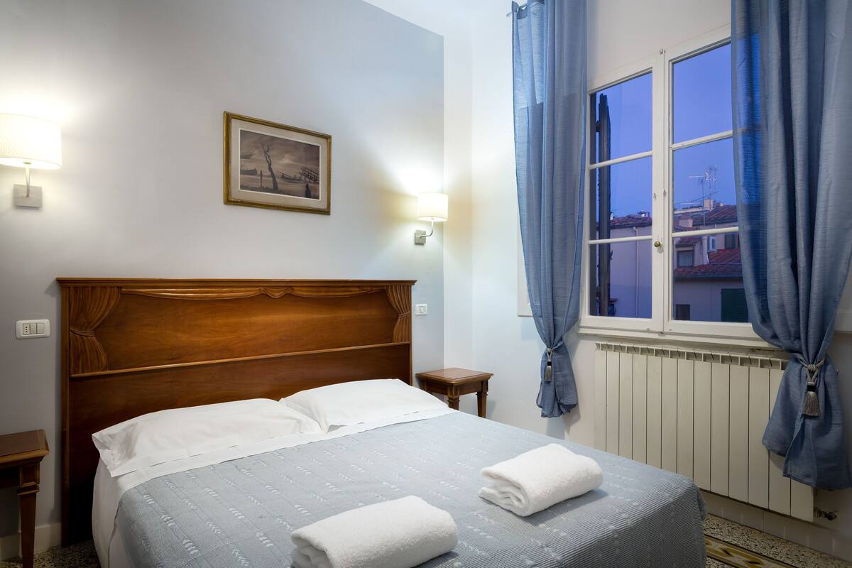 A neatly made bed with white linens and two towels, a wooden headboard, a small bedside table, and a wall-mounted lamp.