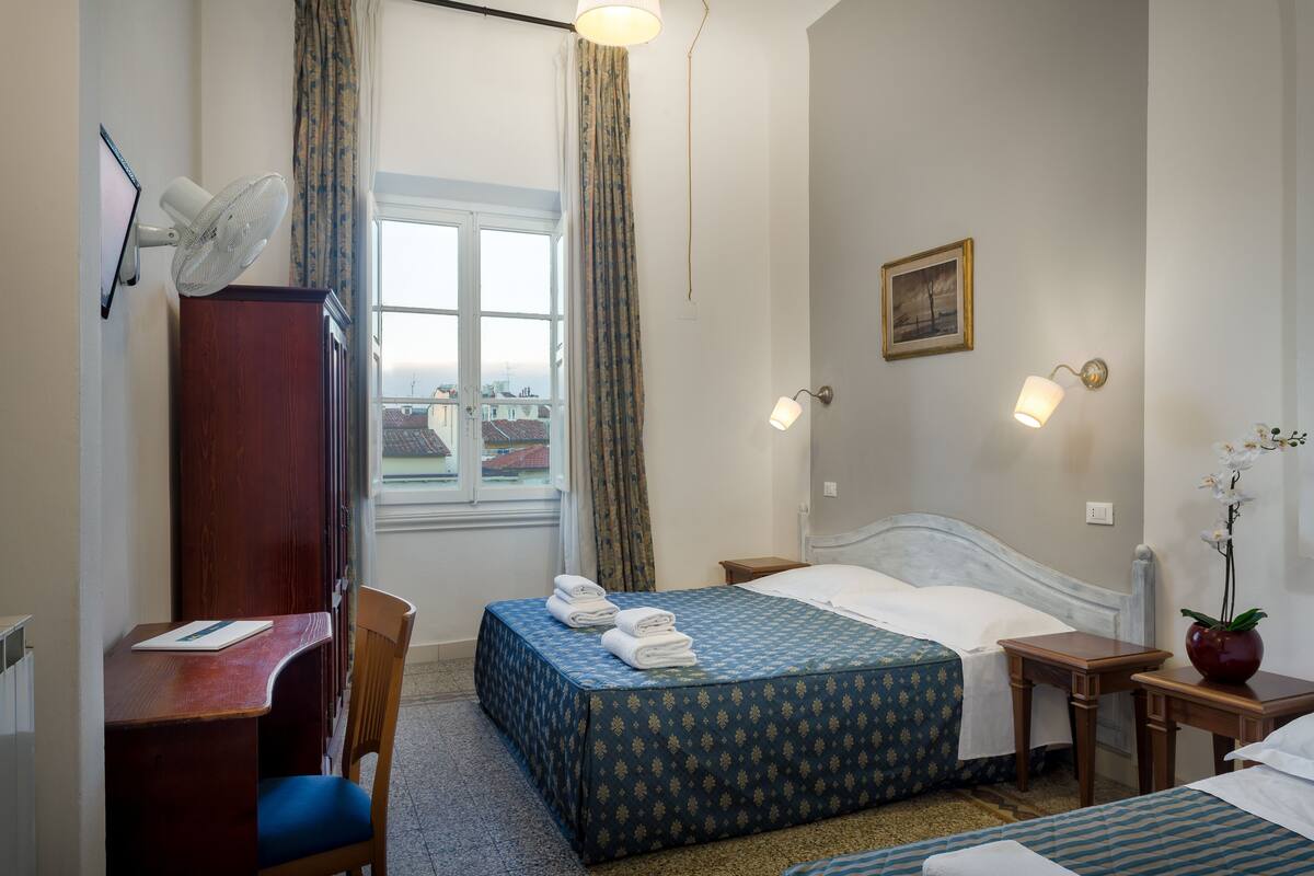 A hotel room with a bed, a desk with a chair, a bedside table, a lamp, a picture on the wall, and a window with curtains.
