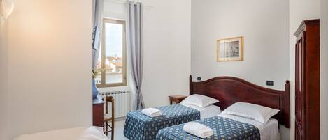 A hotel room with two beds, a wooden headboard, a small bedside table, a chair, a window with curtains, and a picture on the wall.