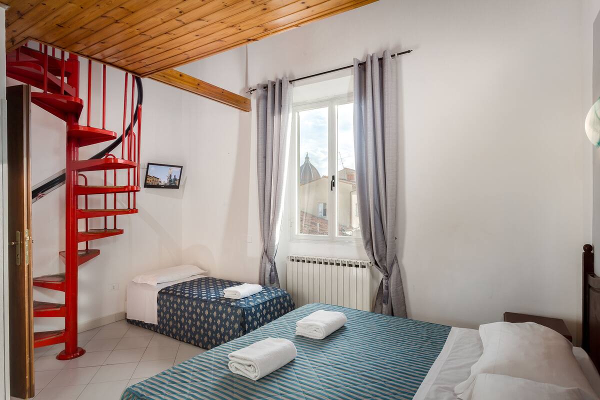A bedroom with a red spiral staircase, two beds with patterned bedding, a window with curtains, and a radiator.