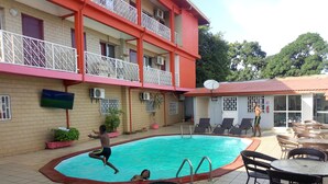 Una piscina con sillas de descanso y mesas, rodeada por un edificio con balcones y plantas en macetas.