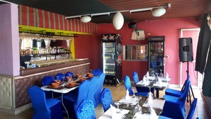 Un restaurante con sillas y mesas tapizadas en azul, una zona de bar con botellas y copas, y una pared roja con techo decorado.