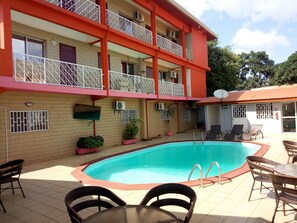A hotel with a swimming pool, outdoor seating, and a red and beige building. - Zambezi Inn Airport Hotel (Conakry)