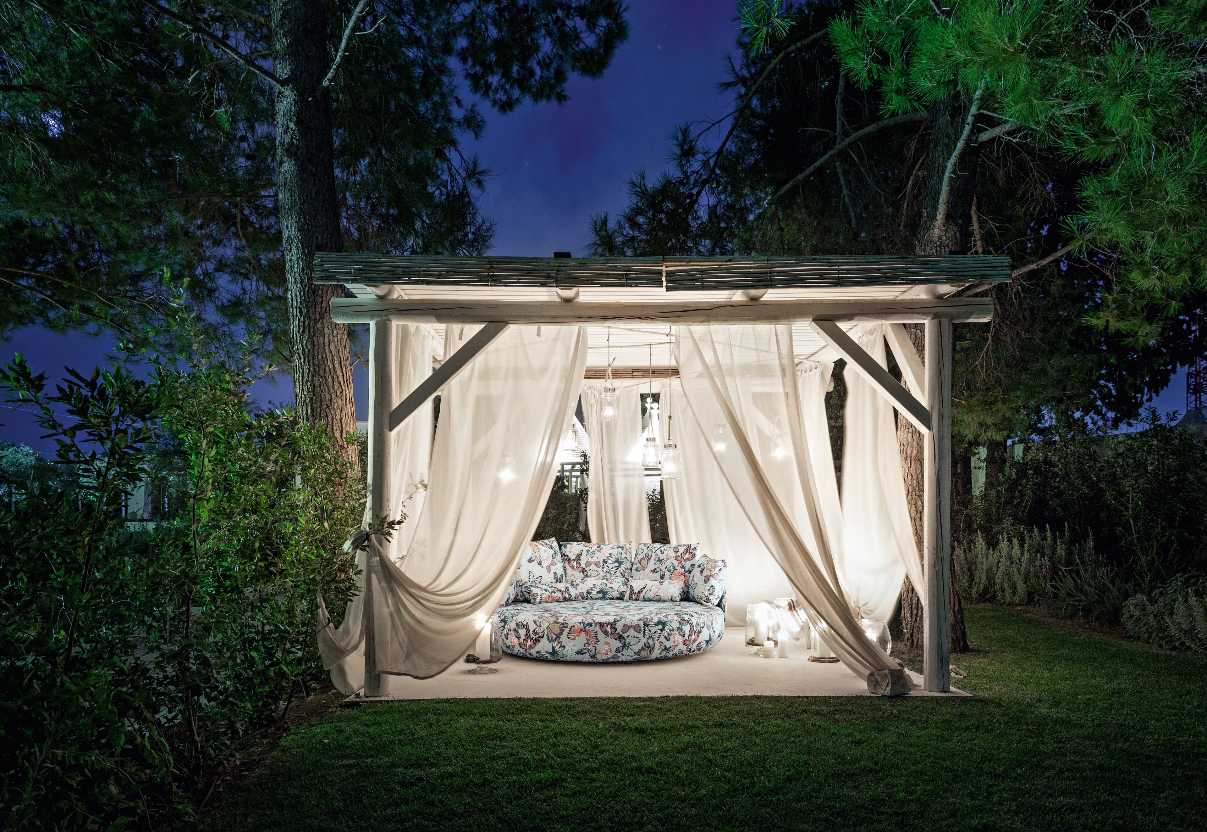 A cozy outdoor seating area with a cushioned bench and a small table, surrounded by draped fabric and trees.