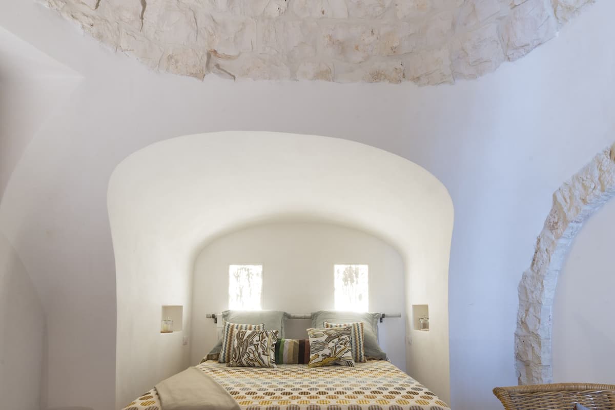 A bedroom with a stone wall, a bed with patterned bedding, a wooden bench, and a wicker chair.