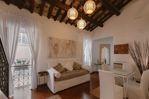 A cozy living room with a sofa, a glass table, and white chairs. There are decorative hanging lights and a view of the outside through a window with curtains.