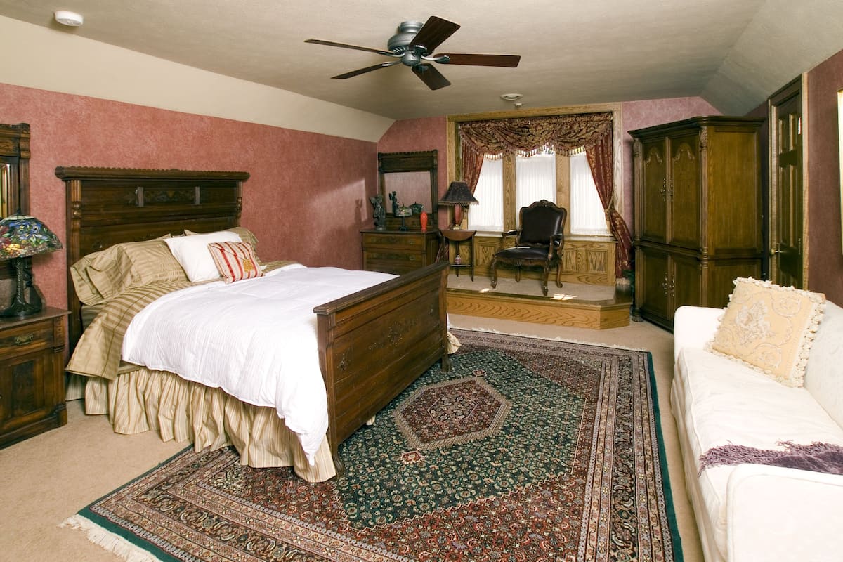 A bedroom with a large bed, a chair, a sofa, a ceiling fan, and a view of a hallway.