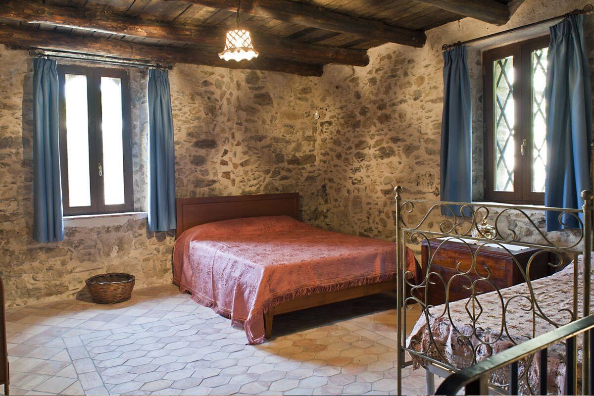 A rustic room with a stone wall, a wooden bed, a metal bed frame, a wooden chest of drawers, and a window with blue curtains.