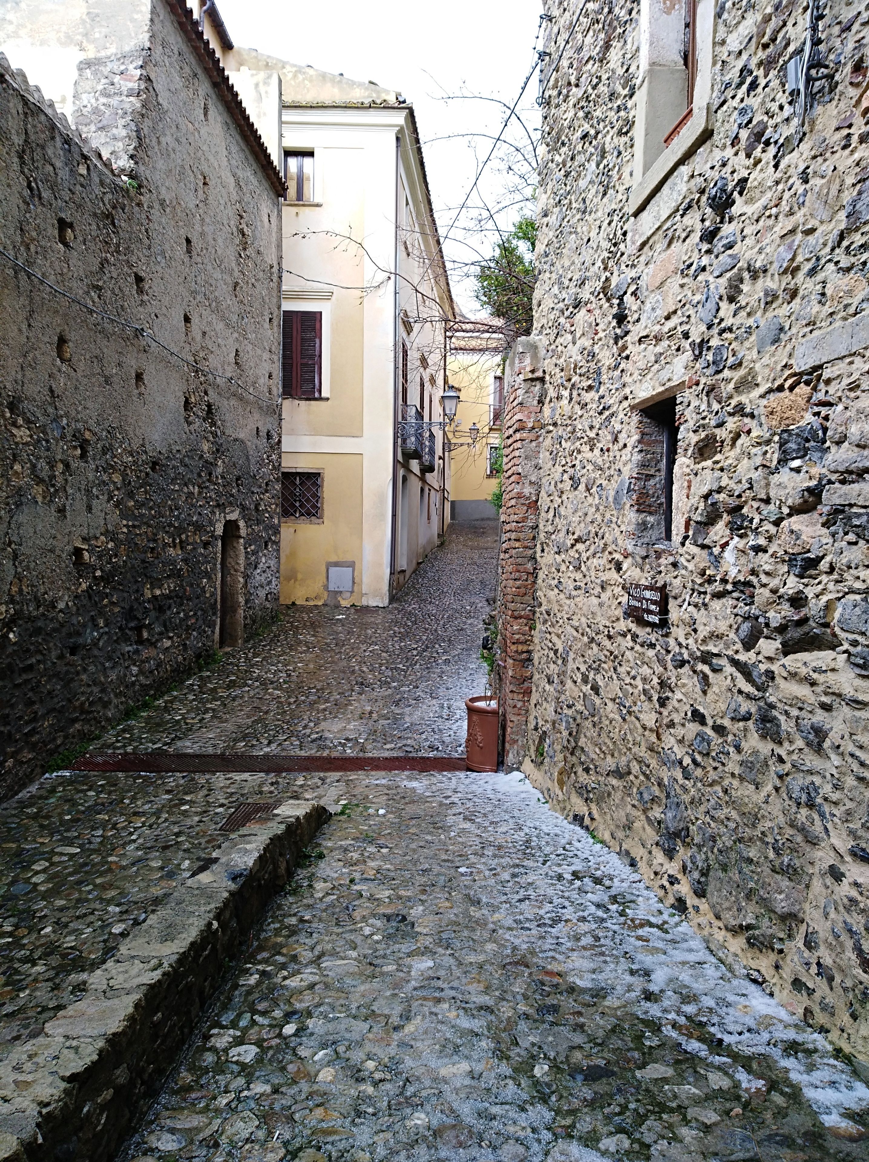 A narrow cobblestone street flanked by historic stone buildings with arched windows and a small drainage channel.