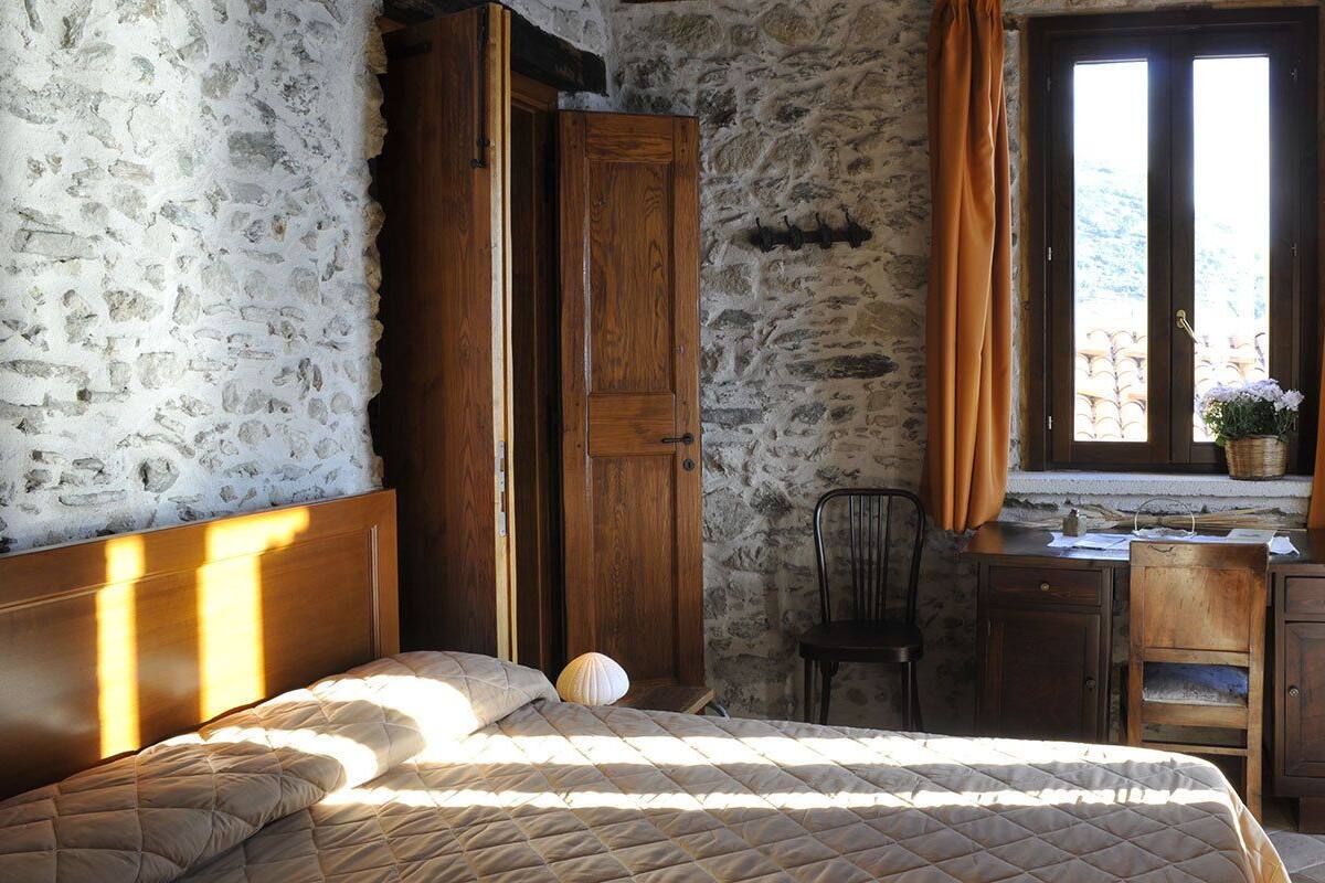 A bedroom with a stone wall, wooden wardrobe, a desk with a chair, and a window with curtains.