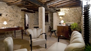 Una habitación rústica con paredes de piedra, vigas de madera y una chimenea de piedra. Hay varias sillas y una mesa con una lámpara.