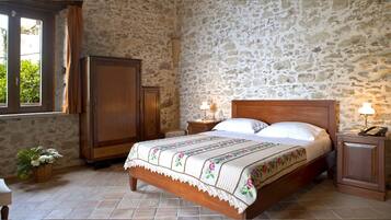 A bedroom with a stone wall, a wooden bed, a nightstand with a lamp, and a wardrobe.