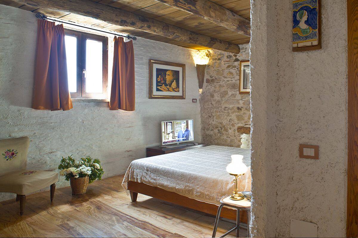 A bedroom with a wooden bed, a bedside table with a lamp, a TV, and a stone wall.