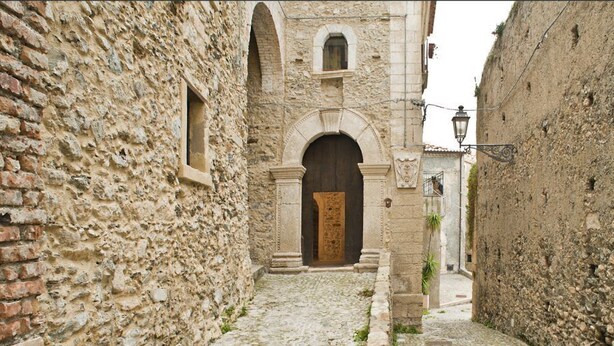 Una estrecha calle empedrada flanqueada por edificios históricos de piedra con arcos y un edificio central con una gran puerta de madera.