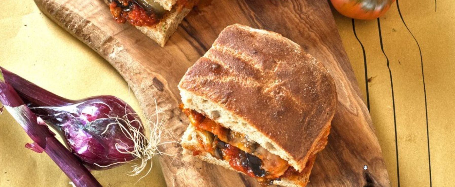 A sandwich on a wooden cutting board with a beetroot and a tomato in the background.