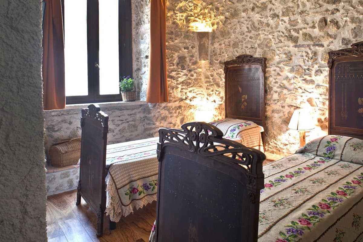 A rustic bedroom with a stone wall, a wooden bed, a nightstand, a dresser, and a window with curtains.